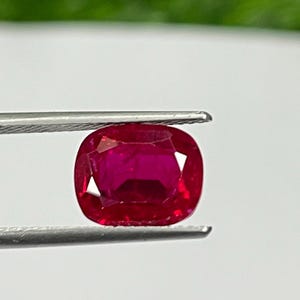 May include: A vibrant, cushion-cut ruby gemstone, held by tweezers against a white and green background. The ruby displays a deep red hue, with a polished surface and rounded corners. The tweezers are silver.