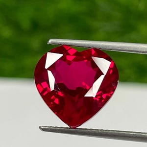 Puede incluir: Una gema roja oscura facetada en forma de corazón es sostenida por unas pinzas contra un fondo verde borroso. Las facetas de la gema capturan la luz, creando un efecto brillante. La gema es de un color rojo vibrante y saturado.