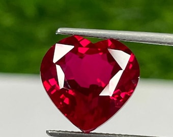 Puede incluir: Una gema roja oscura facetada en forma de corazón es sostenida por unas pinzas contra un fondo verde borroso. Las facetas de la gema capturan la luz, creando un efecto brillante. La gema es de un color rojo vibrante y saturado.