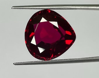 Puede incluir: Una gema facetada de color rojo intenso sujeta con pinzas. La gema tiene forma triangular con un centro en forma de corazón y facetas blancas. El fondo es blanco suave con un toque de verde.