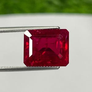 以下が含まれることがあります： ピンセットで保持された、長方形にカットされたルビーの宝石。 宝石は深い赤色で、ファセットが見えます。 背景はぼやけており、緑と白のグラデーションになっています。