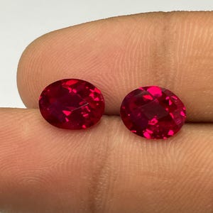 May include: Two oval-cut, faceted, deep red gemstones. The gems are held between fingers against a white and green background. The stones have a brilliant, sparkling appearance, suggesting high clarity and quality. These gems could be used for jewelry.