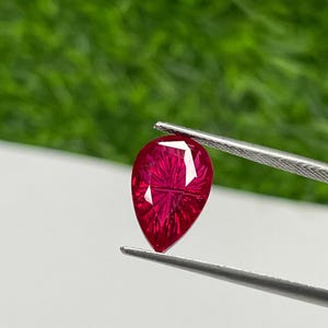 Puede incluir: Una gema facetada, en forma de pera, de color rojo intenso, es sostenida por unas pinzas sobre un fondo blanco y verde. La estructura interna de la gema es visible, reflejando la luz. La gema es de un color vibrante y saturado.