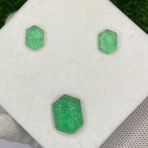 May include: A set of three carved green gemstones, including two earrings and a larger pendant or brooch. Each piece features a floral design within a hexagonal shape, displayed on a white surface against a green background.