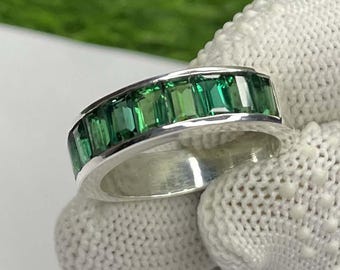 May include: A silver ring with a band of rectangular, emerald-green gemstones. The ring is held by a gloved hand against a white surface, with green foliage in the background. The gemstones are set closely together, creating a continuous line of color.