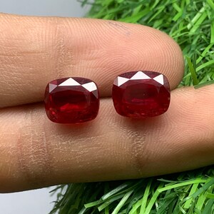 Può includere: Due pietre preziose rosse a cuscino, probabilmente rubini, sono mostrate su un dito. Le pietre preziose sono simili per dimensioni e forma.