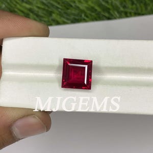 May include: A square-shaped, deep red gemstone with a polished finish. The gemstone is set on a white surface with the text "MJGEMS" printed below it.
