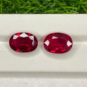 May include: Two oval-shaped, deep red gemstones on a white background. The gemstones are faceted and have a high polish.