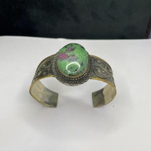 May include: Silver cuff bracelet with an oval, green and red gemstone. The gemstone is set in a decorative, gold-toned bezel. The bracelet has an open design with ornate silver detailing.