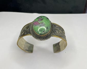 May include: Silver cuff bracelet featuring an oval, green and red gemstone. The gemstone is set in a decorative, gold-toned bezel. The bracelet has an open design with ornate silver detailing.