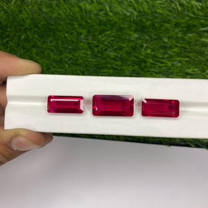 Puede incluir: Tres piedras preciosas rectangulares rojas sobre una superficie blanca. Las piedras preciosas son de diferentes tamaños y están dispuestas en fila.