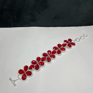 May include: A silver bracelet with a floral pattern, adorned with bright red gemstones. The bracelet is made up of flower-shaped clusters, each with five teardrop-shaped red stones. The clasp is a simple silver toggle.