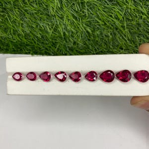 May include: A collection of nine pear-shaped, deep red gemstones displayed on a white surface. The gems vary in size, with a gradient from small to large. The background features green grass.