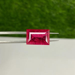 May include: A vibrant red rectangular gemstone with a faceted cut, held in place by a pair of tweezers.