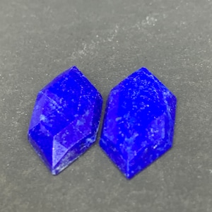 Könnte beinhalten: Zwei tiefblaue, facettierte Edelsteinkabochons. Die Cabochons sind diamantförmig mit abgerundeten Kanten.