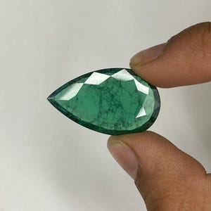 Puede incluir: Una piedra preciosa esmeralda en forma de pera de color verde oscuro. La piedra preciosa tiene un corte facetado y se sostiene en una mano.