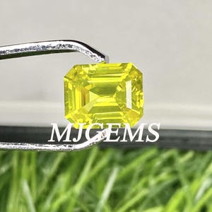 May include: A bright yellow emerald cut gemstone held in tweezers. The gemstone is a vibrant yellow color and has a smooth, polished surface.