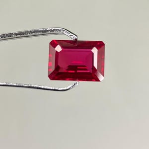 Op de afbeelding: Een levendige rode edelsteen met een rechthoekige smaragd-slijp. De edelsteen wordt vastgehouden door een pincet.