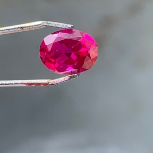 Puede incluir: Una gema ovalada de color rojo intenso y vibrante, sostenida por unas pinzas. La gema está facetada y refleja la luz.