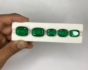 May include: Five emerald-cut green gemstones of varying sizes are displayed on a white surface. The gemstones are arranged in a row, showcasing their vibrant color and clarity. The largest stone is on the left, with the sizes decreasing towards the right.