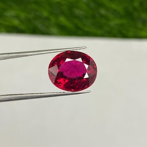 May include: A vibrant red oval gemstone with a faceted cut, held by tweezers against a white background.