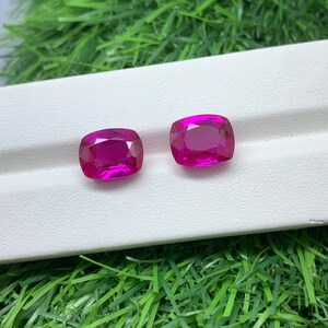 May include: Two vibrant pink gemstones, likely rubies, are displayed on a white surface. The gemstones are cut into a cushion shape and have a polished finish.