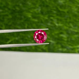 May include: A vibrant, deep red gemstone with a round cut and a faceted surface. The gemstone is held in tweezers against a green background.