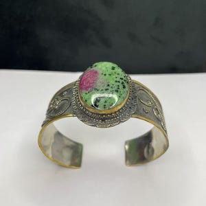 May include: A silver cuff bracelet featuring a large oval gemstone. The gemstone is green with red and black inclusions. The bracelet has a decorative border and gold accents.