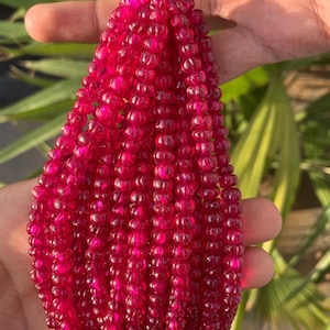 May include: A strand of deep red ruby beads, each bead is faceted and has a slightly rounded shape. The beads are strung on a red thread.