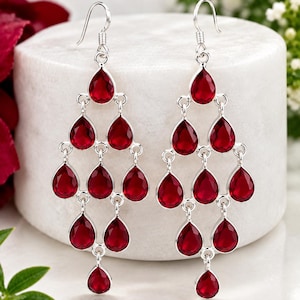 May include: A pair of silver chandelier earrings featuring multiple teardrop-shaped red gemstones arranged in a tiered, cascading design. The earrings hang from silver hooks and include the text MJGEMSIN at the top left corner of the image.