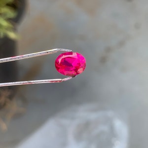 Può includere: Una gemma ovale di colore rosa vibrante tenuta da una pinza. La gemma è un rubino, una pietra preziosa nota per il suo colore rosso intenso.