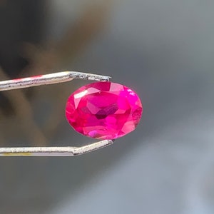 Könnte beinhalten: Ein leuchtend rosa ovaler Edelstein, der mit einer Pinzette gehalten wird. Der Edelstein ist facettiert und reflektiert Licht.