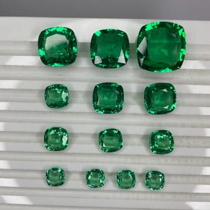May include: An assortment of emerald-cut green gemstones of varying sizes. The gems are arranged in rows, showcasing their vibrant color and facets. The largest gem is at the top right, with smaller ones descending in size.