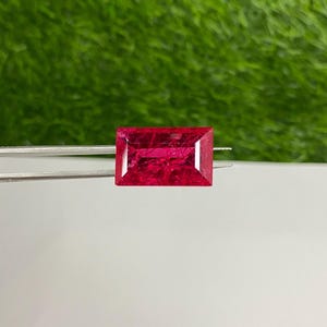 Op de afbeelding: Een levendige rode edelsteen met een rechthoekige snede, vastgehouden in een pincet tegen een groene achtergrond.