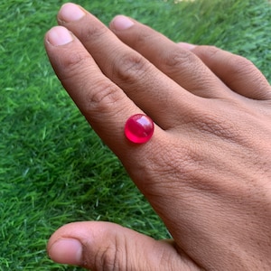 Puede incluir: Una piedra preciosa redonda de color rojo brillante, probablemente un rubí, se muestra en el dedo de una persona.