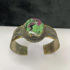 May include: A silver cuff bracelet featuring an oval gemstone with green, pink, and black hues. The bracelet has detailed scrollwork and gold highlights. The cuff is open at the base. The gemstone is approximately 2.5 cm in length.