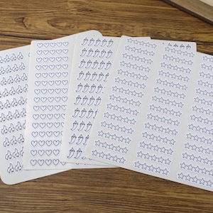 May include: A collection of white sticker sheets with various blue outline designs, including flames, hearts, chili peppers, and stars. The stickers are arranged on a wooden surface, offering a variety of decorative options.