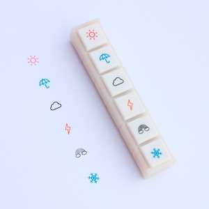 May include: A white plastic stamp set with six different weather icons: sun, umbrella, cloud, lightning bolt, rainbow, and snowflake. The icons are in different colors, including red, blue, orange, and black.