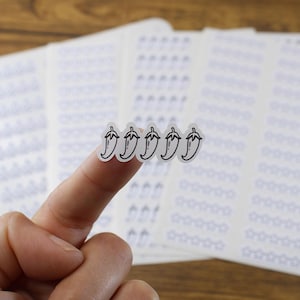 Puede incluir: Una mano sostiene cinco pegatinas pequeñas y transparentes con el contorno negro de un chile. Las pegatinas están sobre una hoja de papel blanco con un patrón de pequeñas estrellas. El fondo es una superficie de madera.