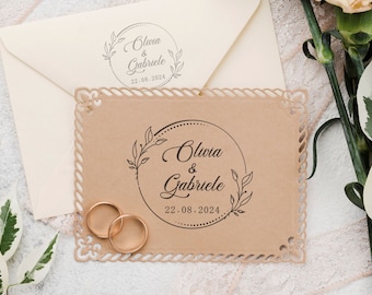 Könnte beinhalten: Eine braune Karte mit einem dekorativen Rand und einem runden Stempel mit dem Text "Olivia & Gabriele 22.08.2024" in einem Kranz aus Blättern. Zwei goldene Eheringe liegen auf der Karte.