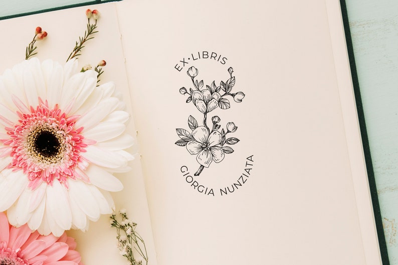 May include: A white book with a black and white floral design stamp on the front cover. The stamp says "Ex Libris Giorgia Nunziata".
