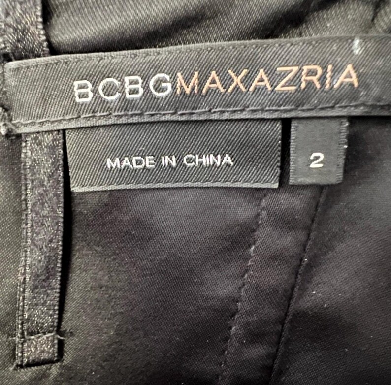 May include: Close-up of a black garment with a label that reads "BCBGMAXAZRIA" in white and gold lettering. Below the brand name, the label states "MADE IN CHINA." A separate tag indicates the size is "2."