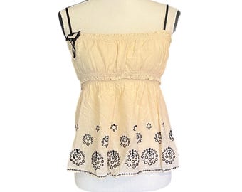 May include: A cream-colored camisole top with black embroidered floral designs. The top features thin black straps and a decorative bow. The waistline has a ruffled detail. The bottom hemline has a scalloped edge.