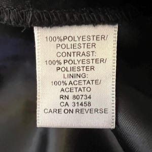 May include: A white clothing tag with black text. The tag lists fabric composition: 100% polyester, 100% acetate. It also includes the RN and CA numbers. The tag is attached to a black fabric.