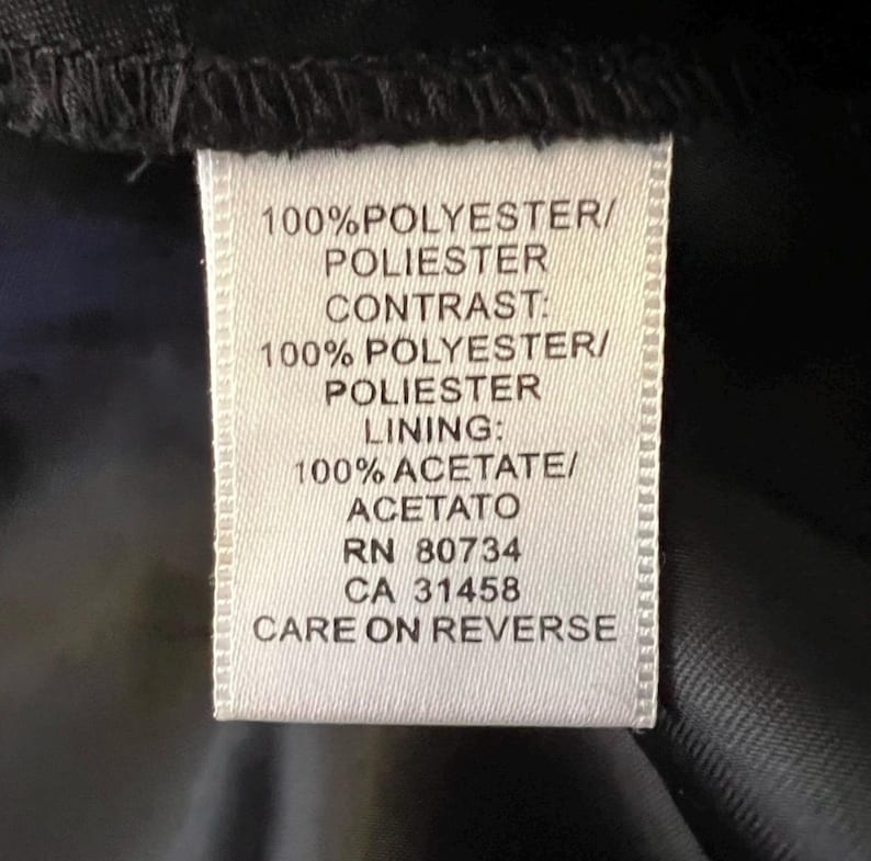 May include: A white clothing tag with black text. The tag lists fabric composition: 100% polyester, 100% acetate. It also includes the RN and CA numbers. The tag is attached to a black fabric.