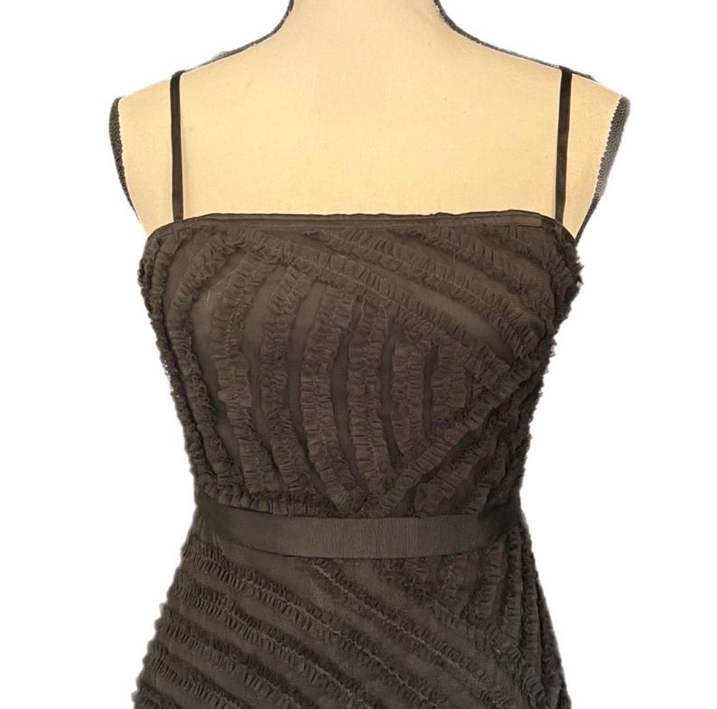 May include: A dark brown, strapless dress with a textured, ruffled design. The dress features thin spaghetti straps and a matching belt around the waist. The fabric has a layered, diagonal pattern.