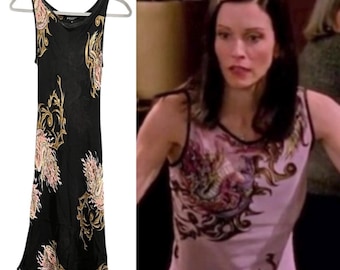 May include: Black sleeveless dress with a floral pattern in shades of pink, gold, and cream. The dress has a scoop neckline and a flowing silhouette. The dress is hanging on a gray hanger.