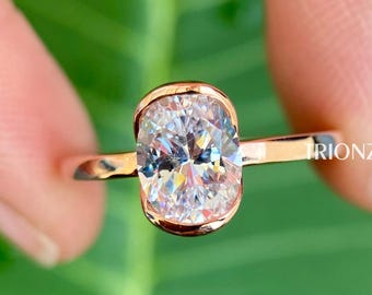 May include: A rose gold engagement ring with an oval-shaped center stone. The ring is set on a thin band and features a delicate, curved design. The ring is displayed on a green leaf.