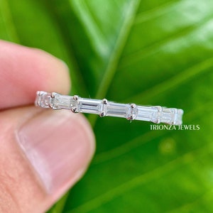 May include: A silver band ring with a row of clear baguette-shaped diamonds. The ring is on a finger against a green leafy background. The text "TRIONZA JEWELS" is visible in the lower right corner.