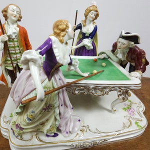 May include: A porcelain figurine set depicting four people playing billiards. The figures are dressed in 18th-century clothing and are posed around a green felt billiard table. The table has ornate gold trim and sits on a white base with floral details.